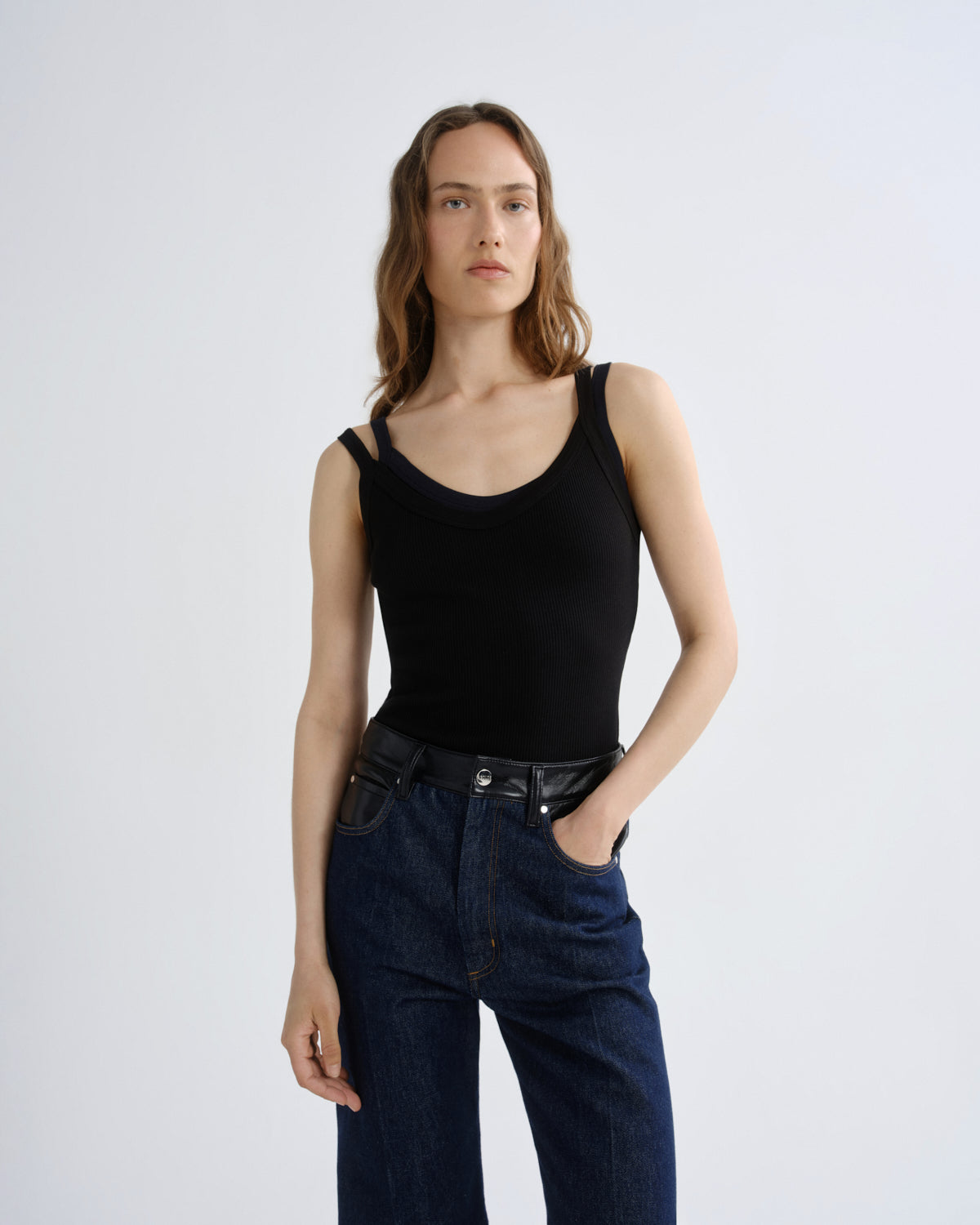 A woman modeling a black ribbed tank top with shoulder cutouts and dark blue jeans against a light gray studio background.