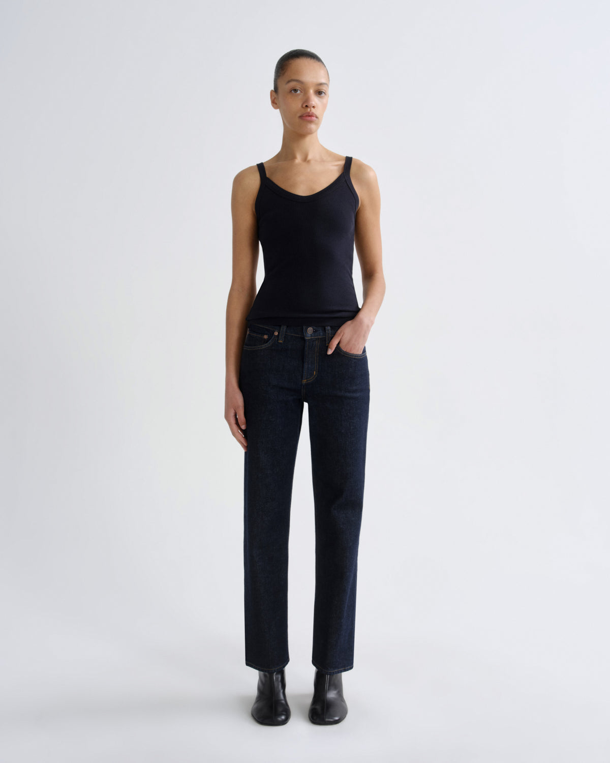 A model stands against a light gray studio background wearing a black ribbed camisole tank top, dark blue straight-leg jeans, and black boots.