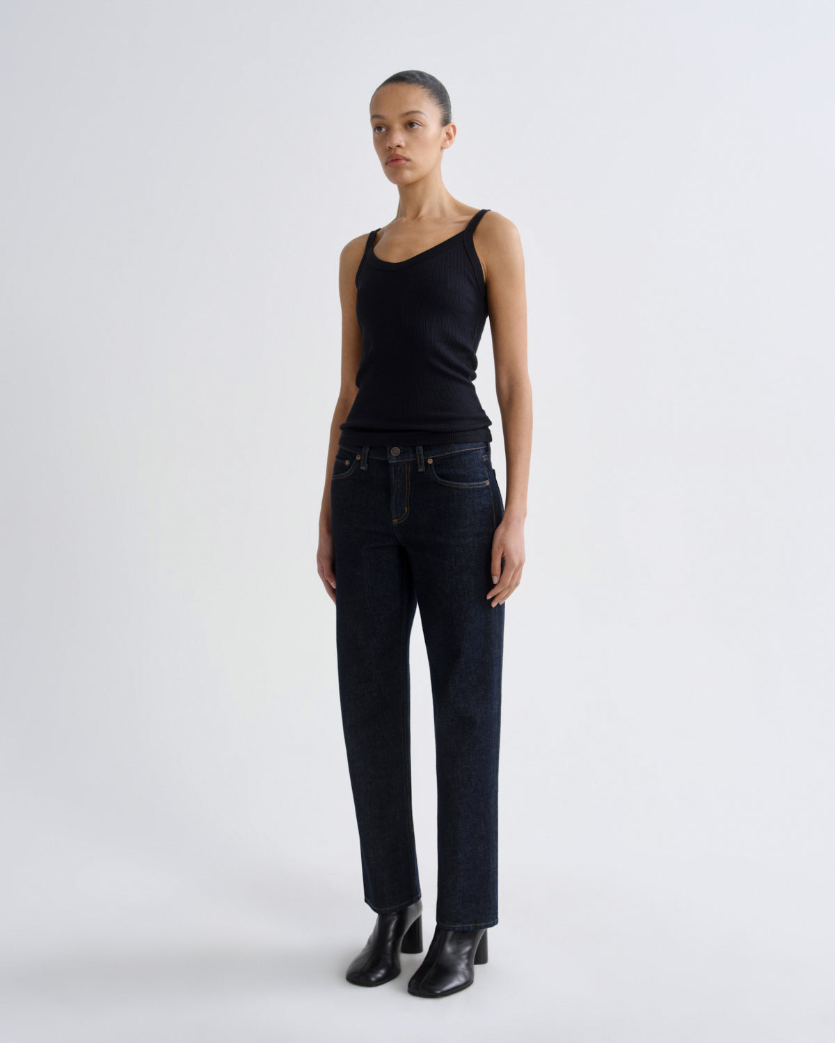 A model stands against a light gray studio background wearing a black ribbed tank top, dark blue straight-leg jeans, and black leather ankle boots.