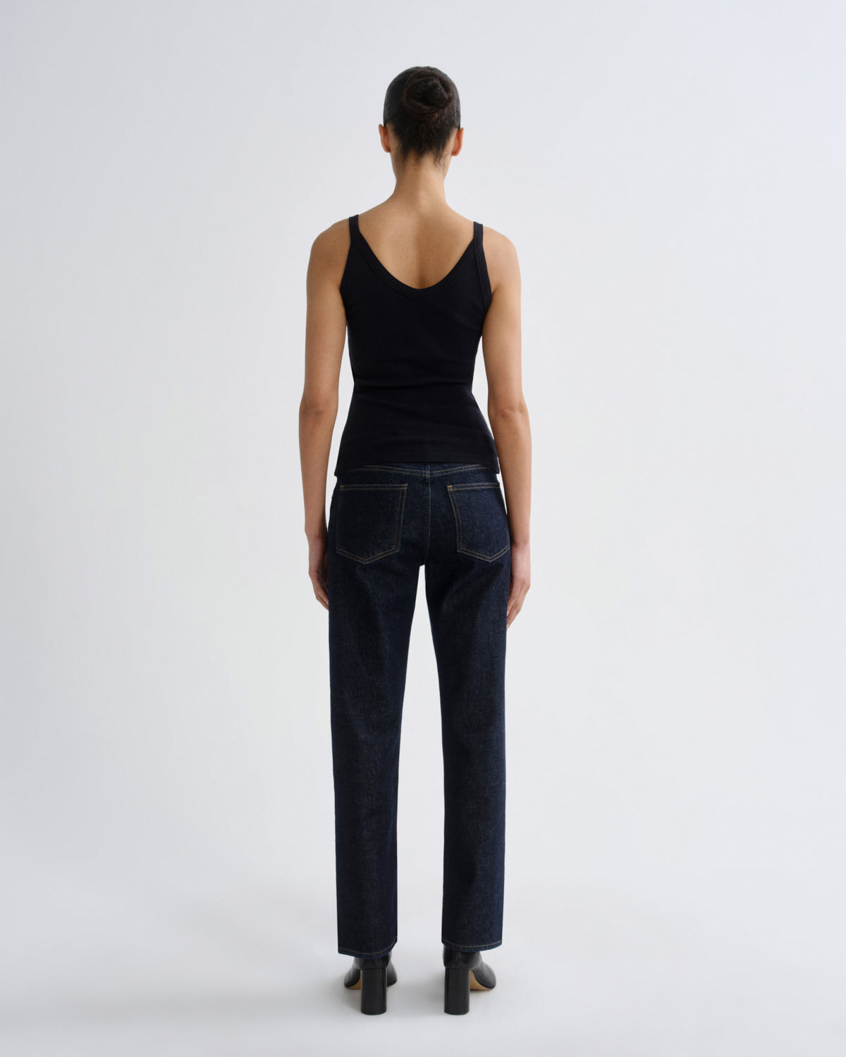 A model viewed from the back wearing a black tank top and dark blue straight-leg jeans, standing against a light gray studio background.