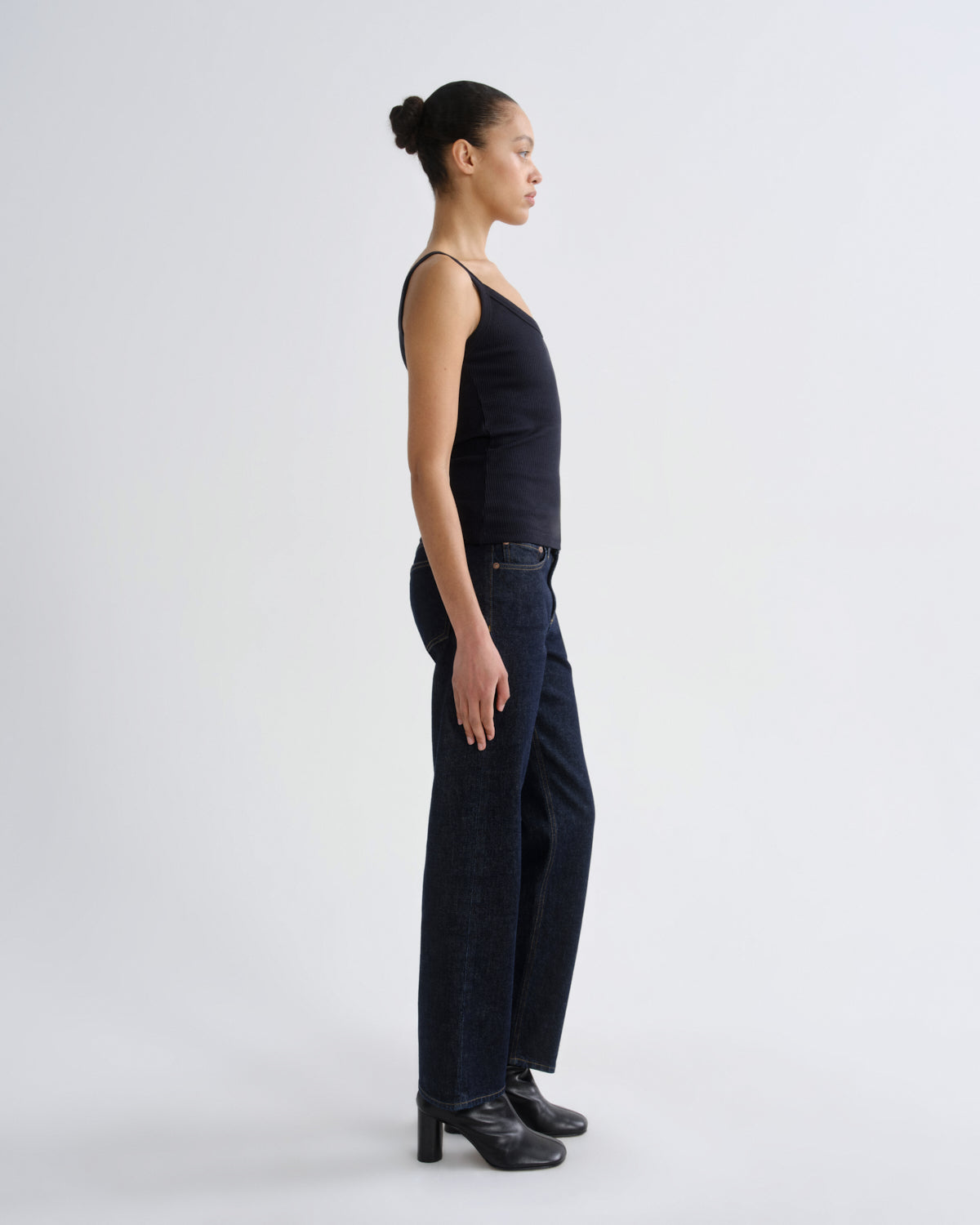 A model in profile wearing a black sleeveless top, dark wide-leg jeans, and black heeled boots, standing against a plain light gray studio background.