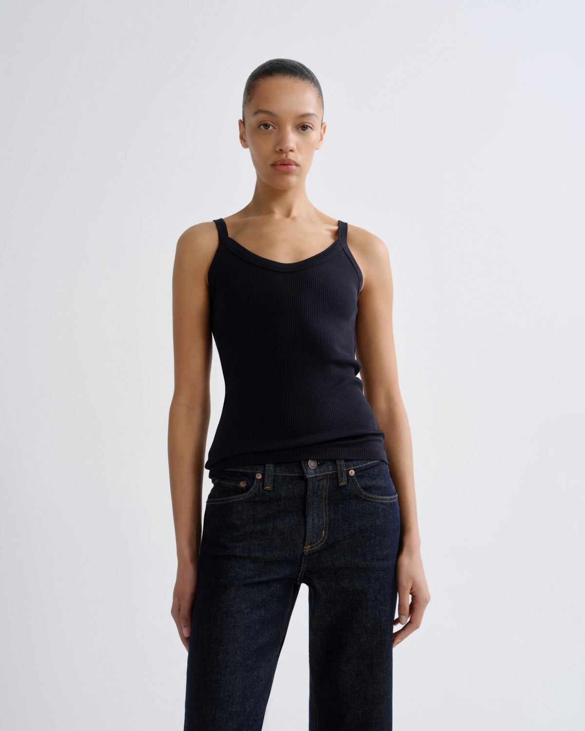 A model wearing a black ribbed knit camisole tank top and dark blue jeans, standing against a light gray studio background.