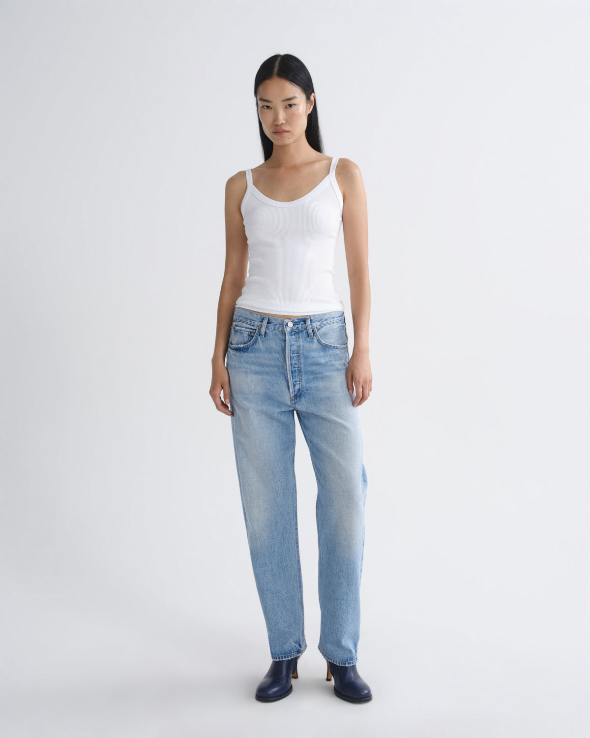 A model stands against a light gray studio background wearing a white ribbed camisole tank top, light-wash blue straight-leg jeans, and dark navy heeled boots.