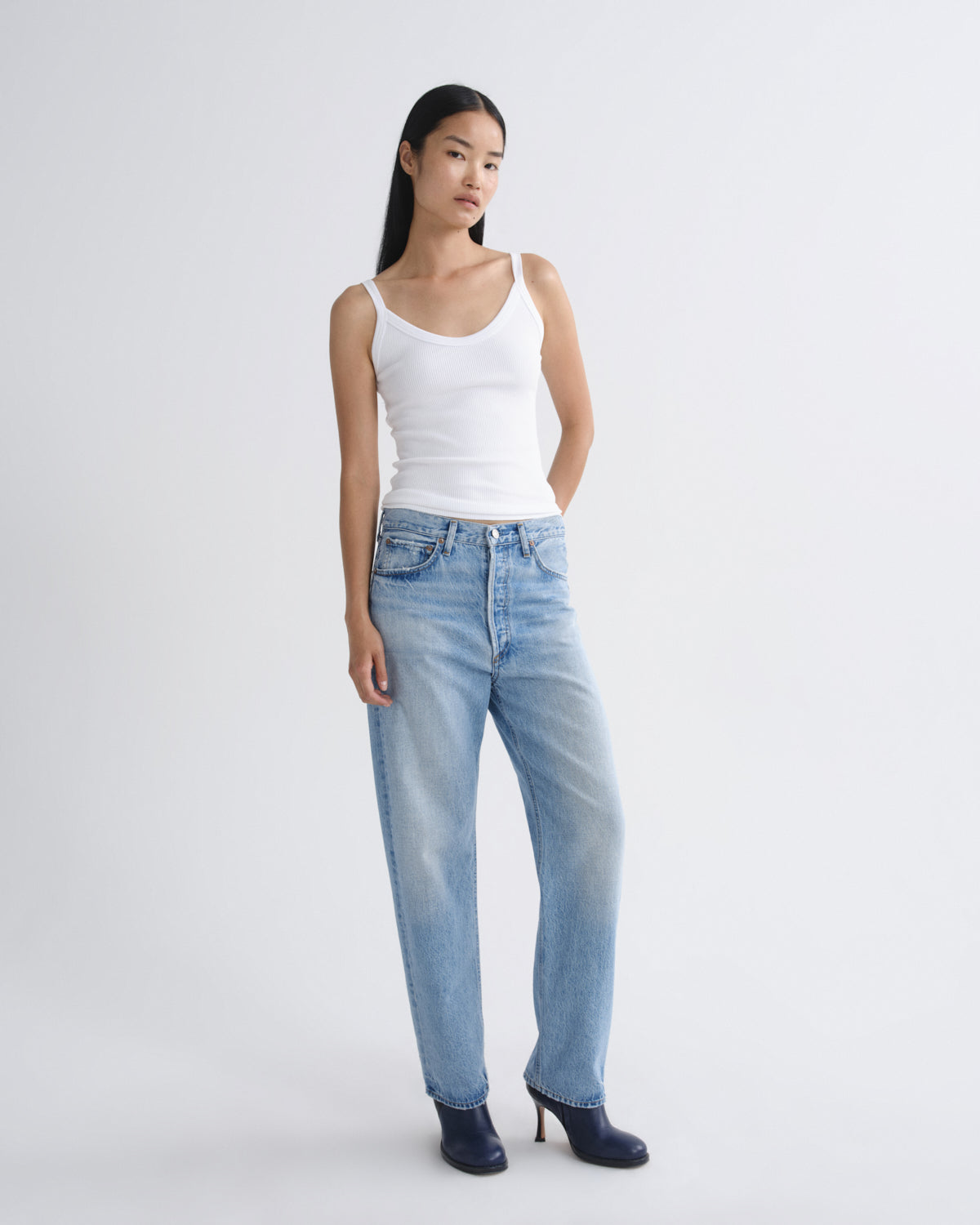 A model wearing a white ribbed tank top, light-wash blue straight-leg jeans, and dark navy heeled ankle boots, standing against a light gray studio background.