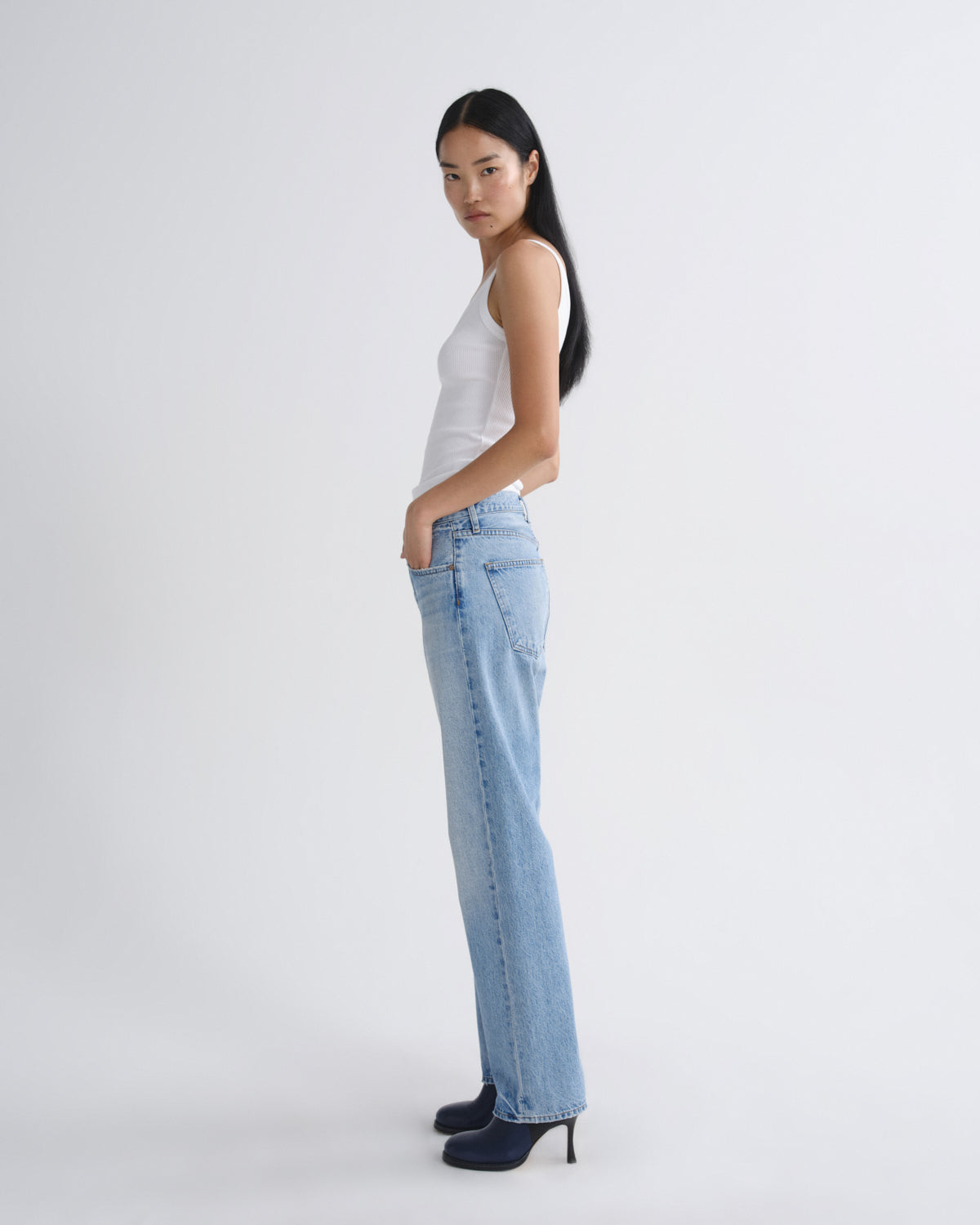 A woman modeling light-wash blue straight-leg jeans, paired with a white tank top and dark navy heels, standing in profile against a plain light gray studio background.