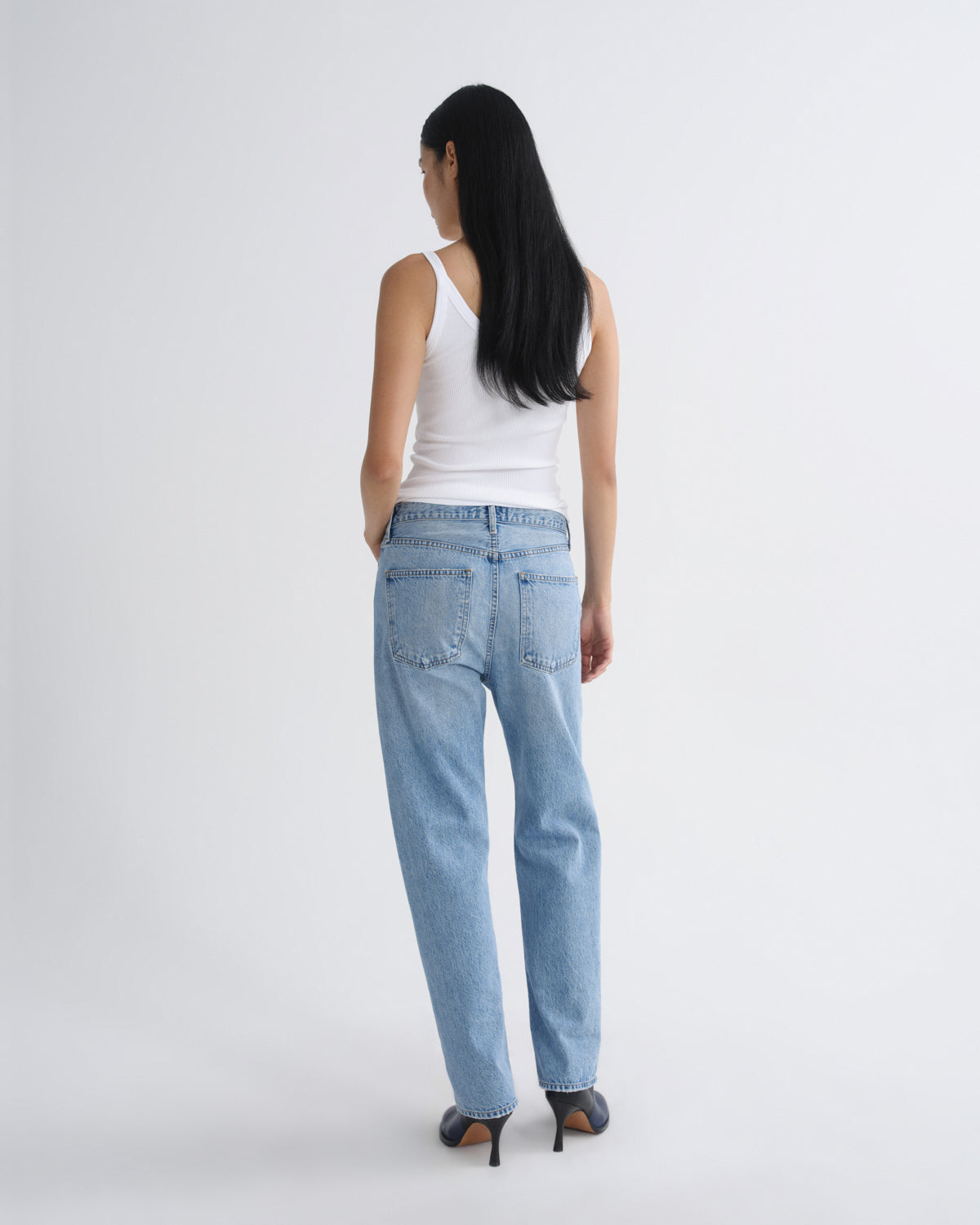 A woman is shown from behind wearing light-wash blue jeans, a white tank top, and black high heels against a plain light gray studio background.