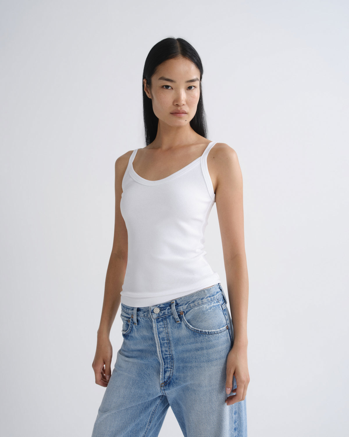 A model wearing a white ribbed tank top and light blue jeans, standing against a plain light gray studio background.