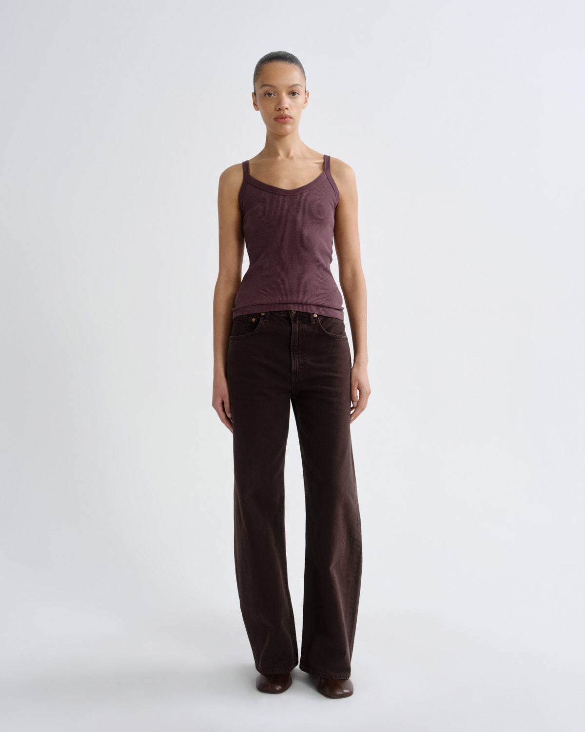 A model stands against a light gray studio background wearing a sleeveless mauve-brown ribbed tank top and dark brown wide-leg jeans.
