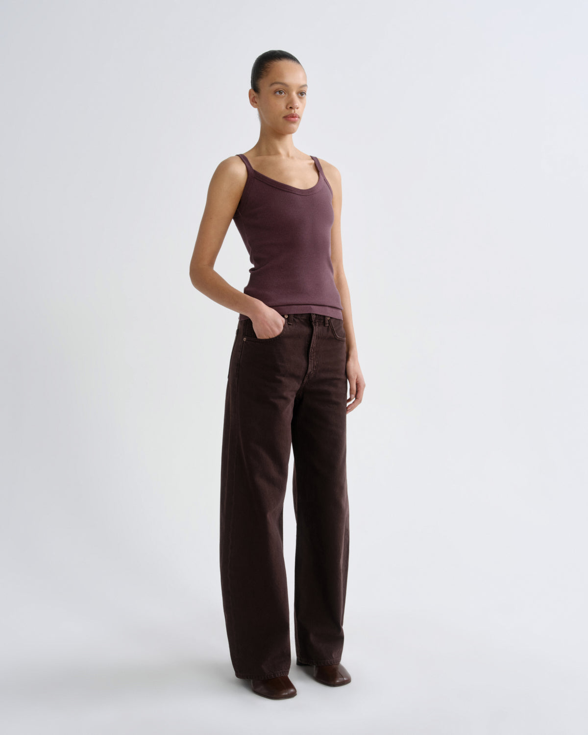 A model stands in a studio wearing a sleeveless dark plum tank top and dark brown wide-leg trousers against a light gray background.