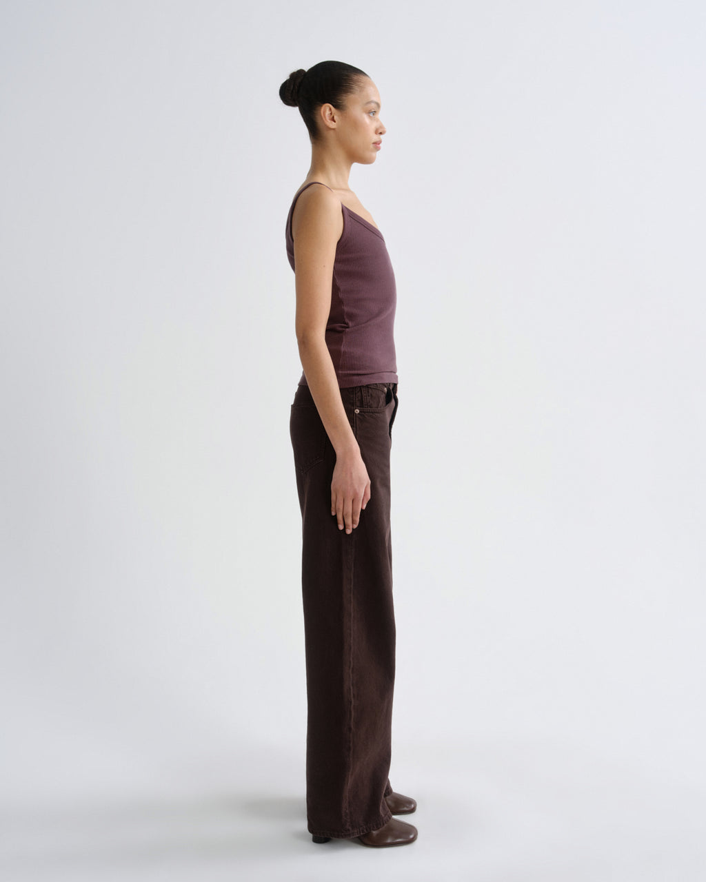 A model in profile wearing a sleeveless mauve-brown top and dark brown wide-leg pants, standing against a light gray studio background.
