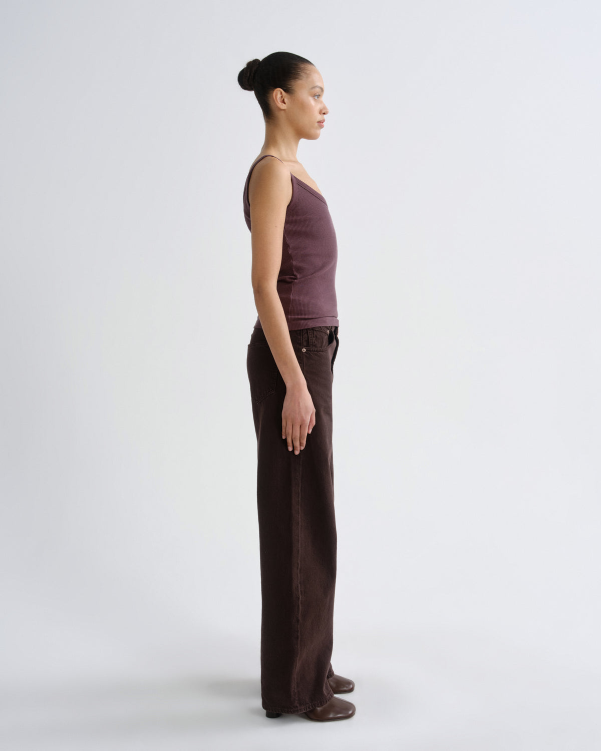 A model in profile wearing a sleeveless mauve-brown top and dark brown wide-leg pants, standing against a light gray studio background.