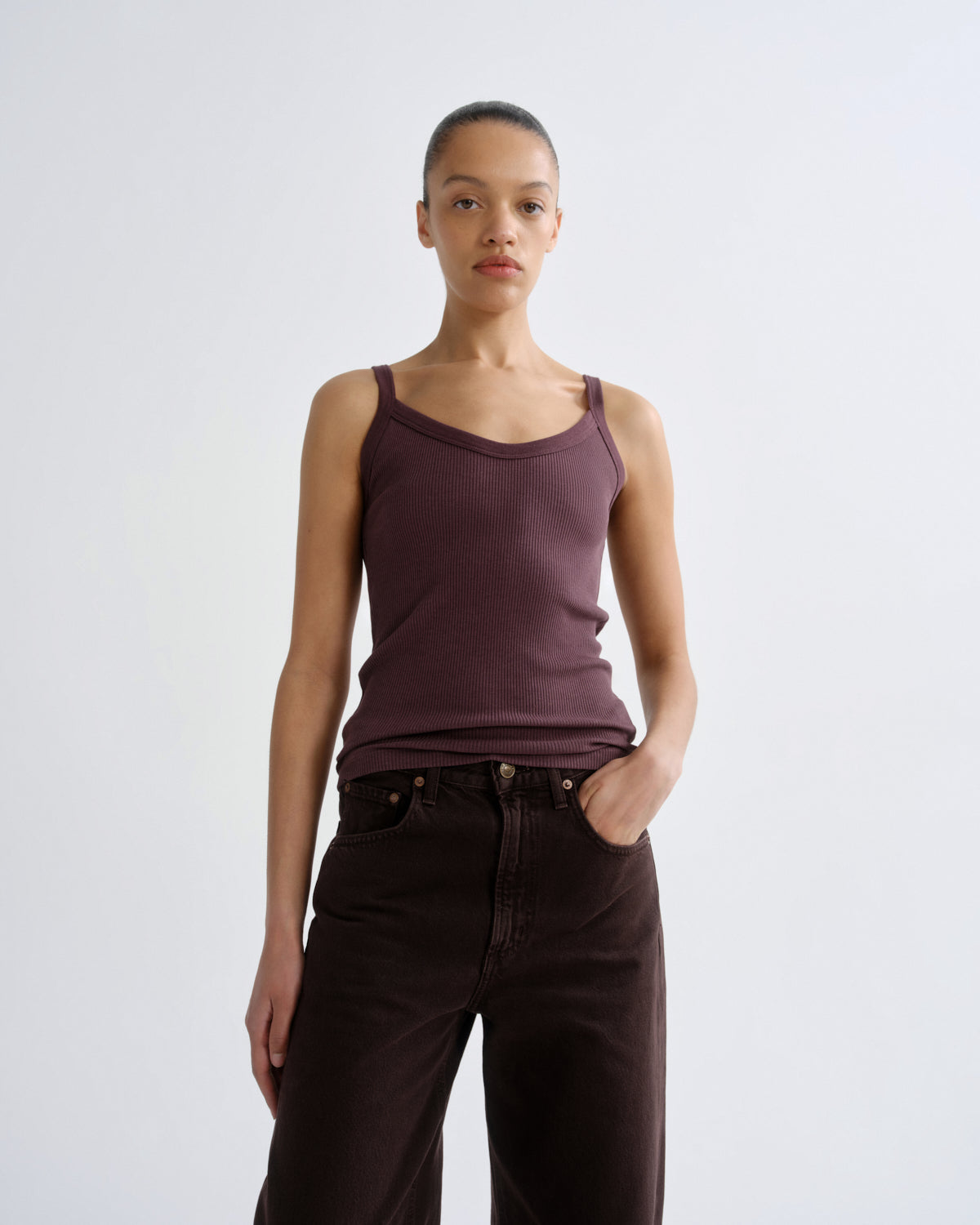 A model wearing a dark plum purple ribbed knit camisole tank top and dark brown corduroy pants, standing against a light gray studio background.