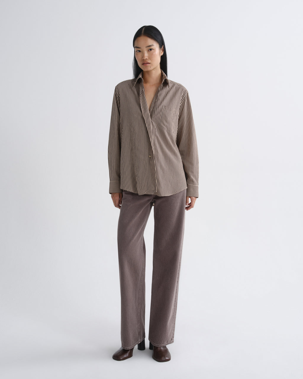 A model stands against a light gray studio background wearing a brown and beige vertically striped button-up shirt with matching brown wide-leg trousers and dark brown shoes.