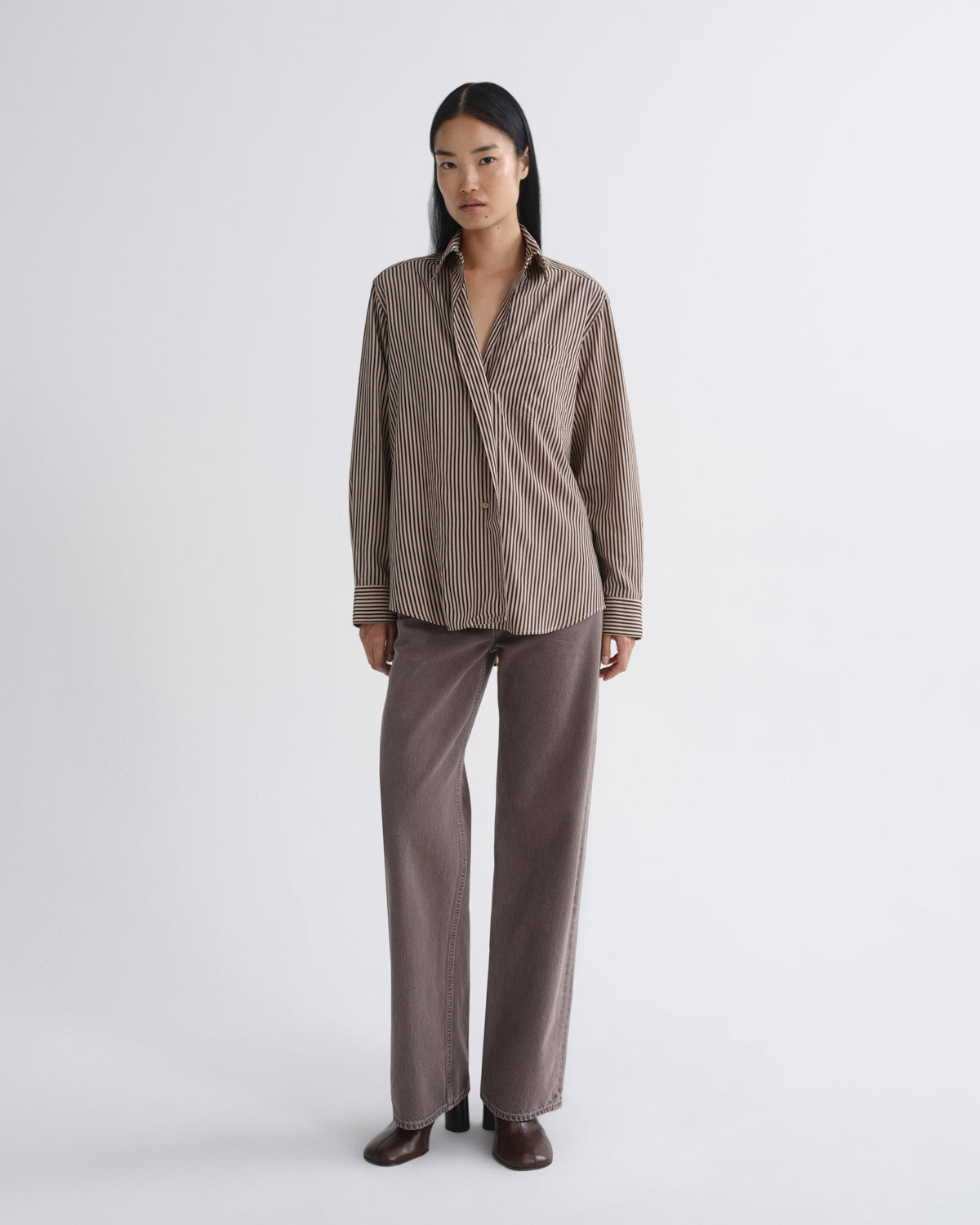 A model stands against a light gray studio background wearing a brown and beige vertically striped button-up shirt with matching brown wide-leg trousers and dark brown shoes.