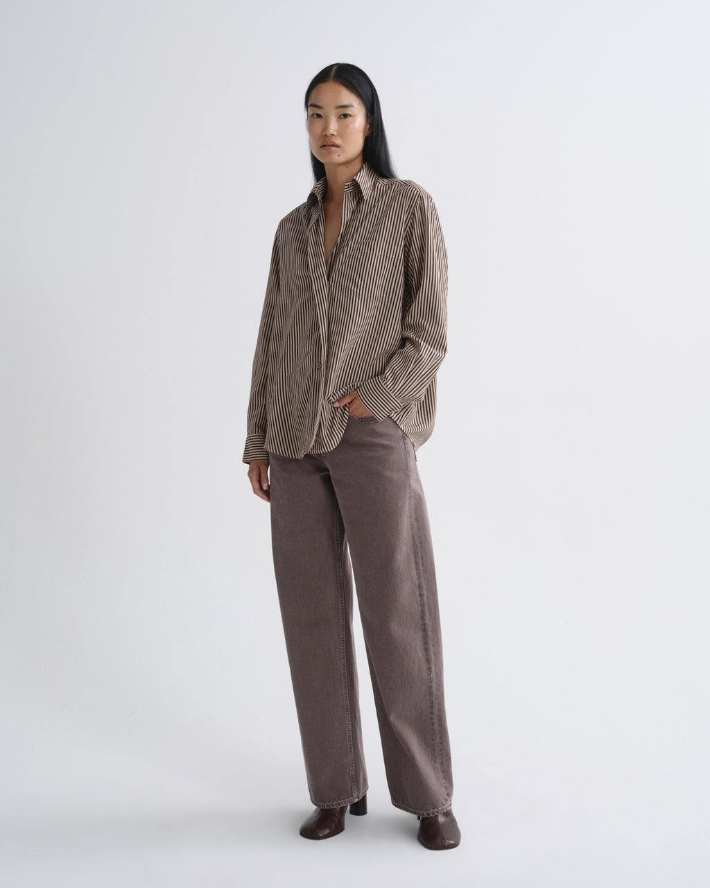 A model stands against a light gray studio background wearing a brown and beige vertically striped button-down shirt and matching brown wide-leg trousers.