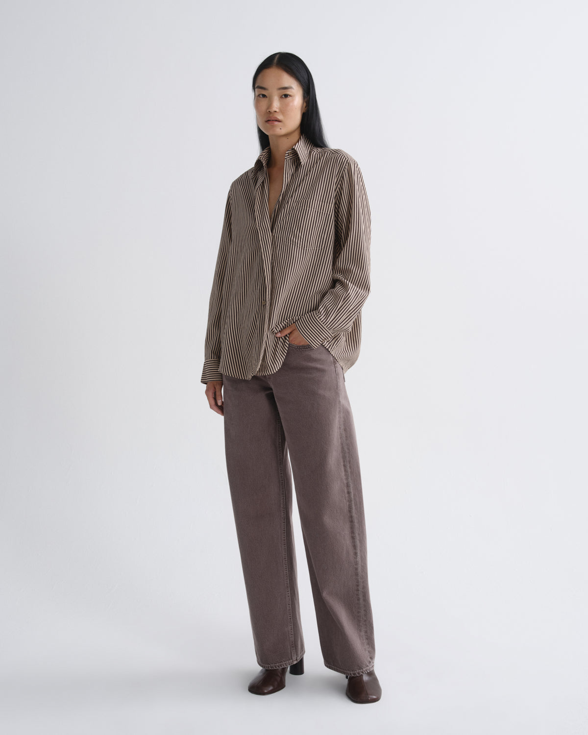 A model stands against a light gray studio background wearing a brown and beige vertically striped button-down shirt and matching brown wide-leg trousers.