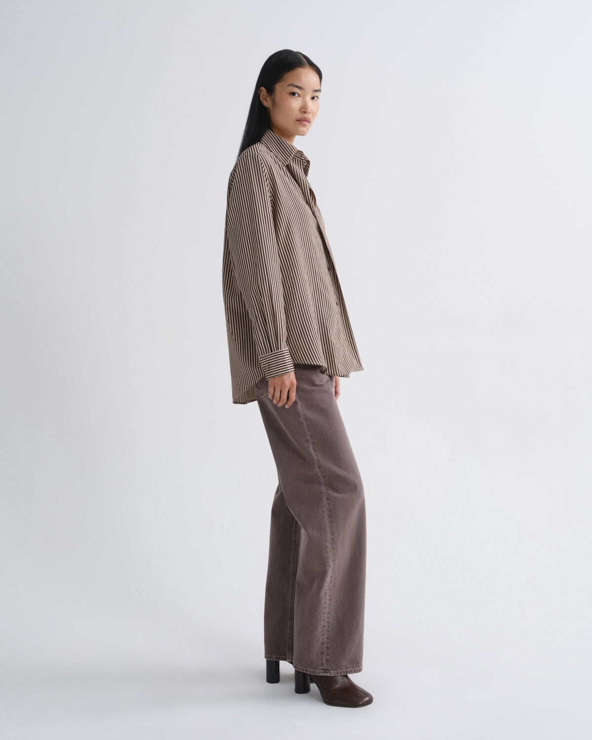 A model is shown from the side wearing a brown and beige vertically striped button-up shirt and matching brown wide-leg trousers, standing against a plain light gray studio background.