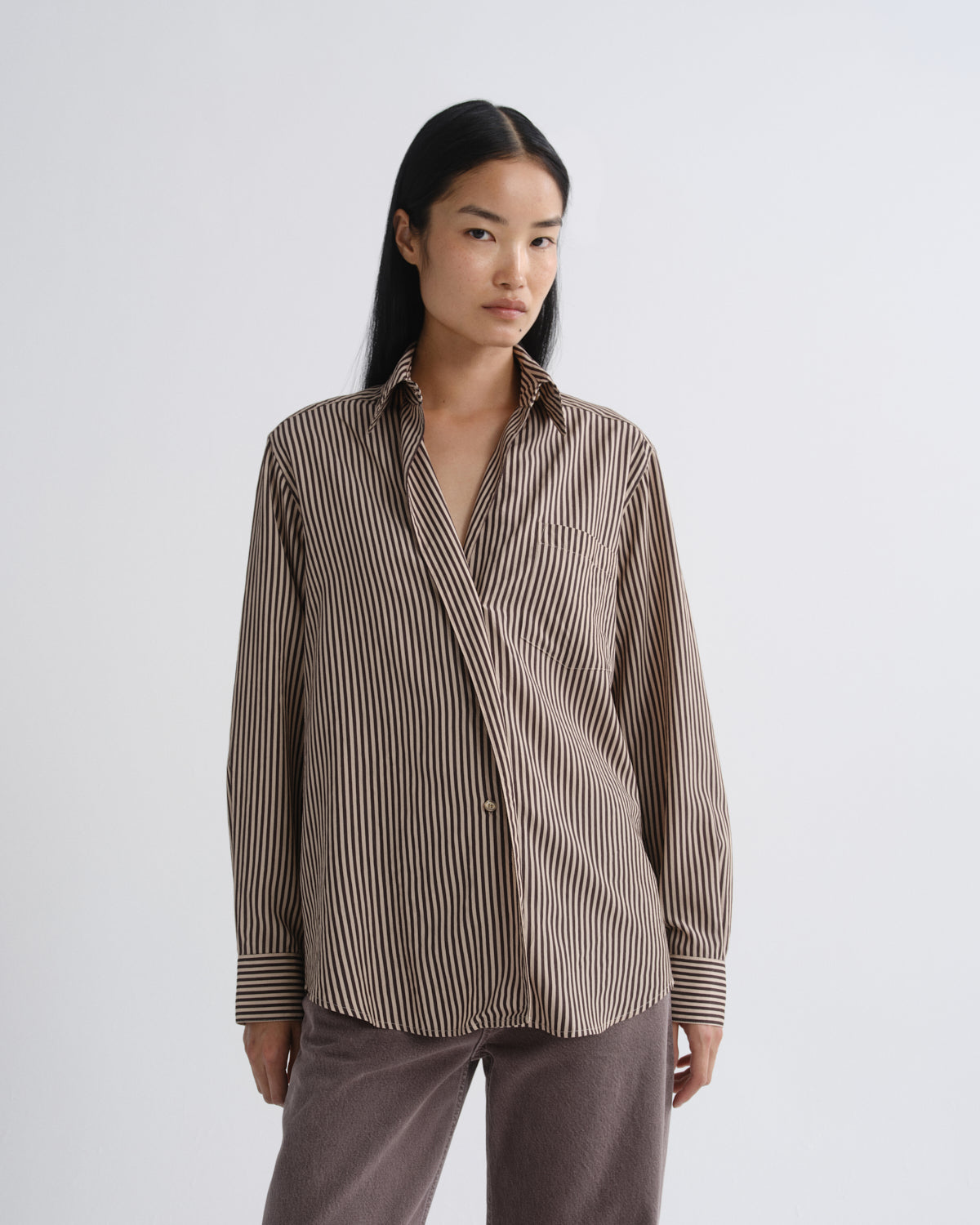 A woman modeling a brown and beige vertically striped button-up shirt with a relaxed fit, paired with brown pants, against a light gray studio background.