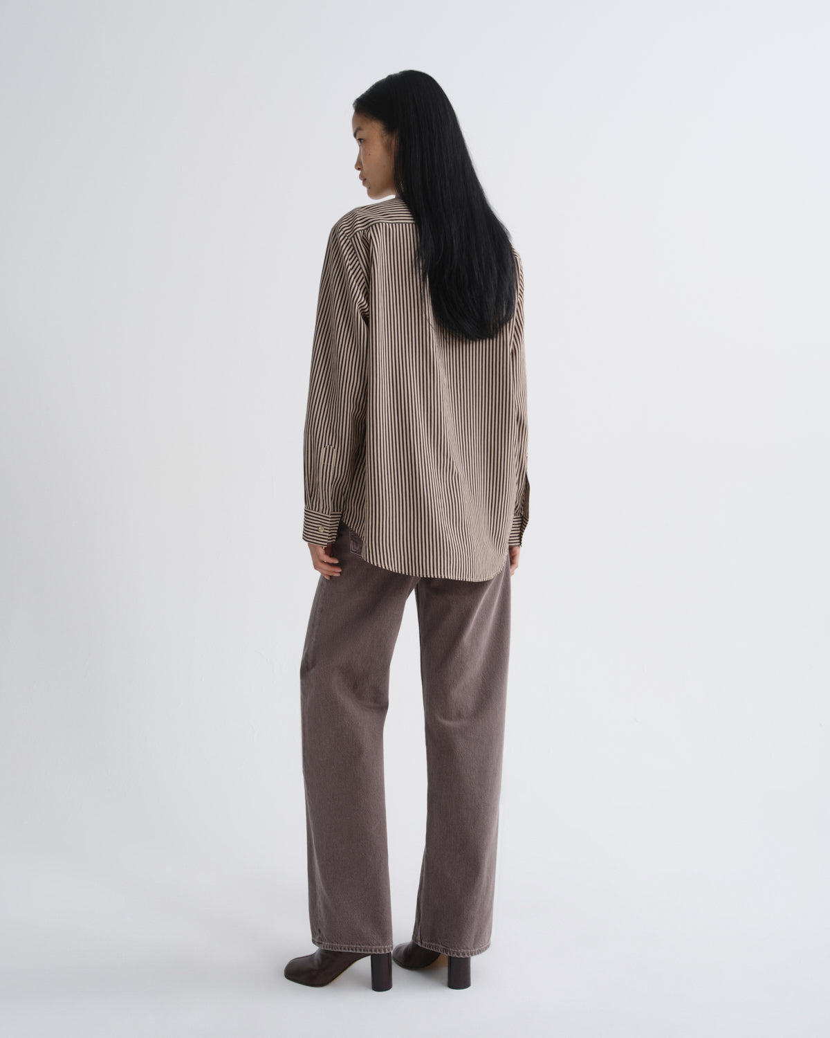 A woman is shown from behind wearing a beige and brown vertically striped long-sleeve shirt and wide-leg taupe trousers, standing against a plain light gray studio background.