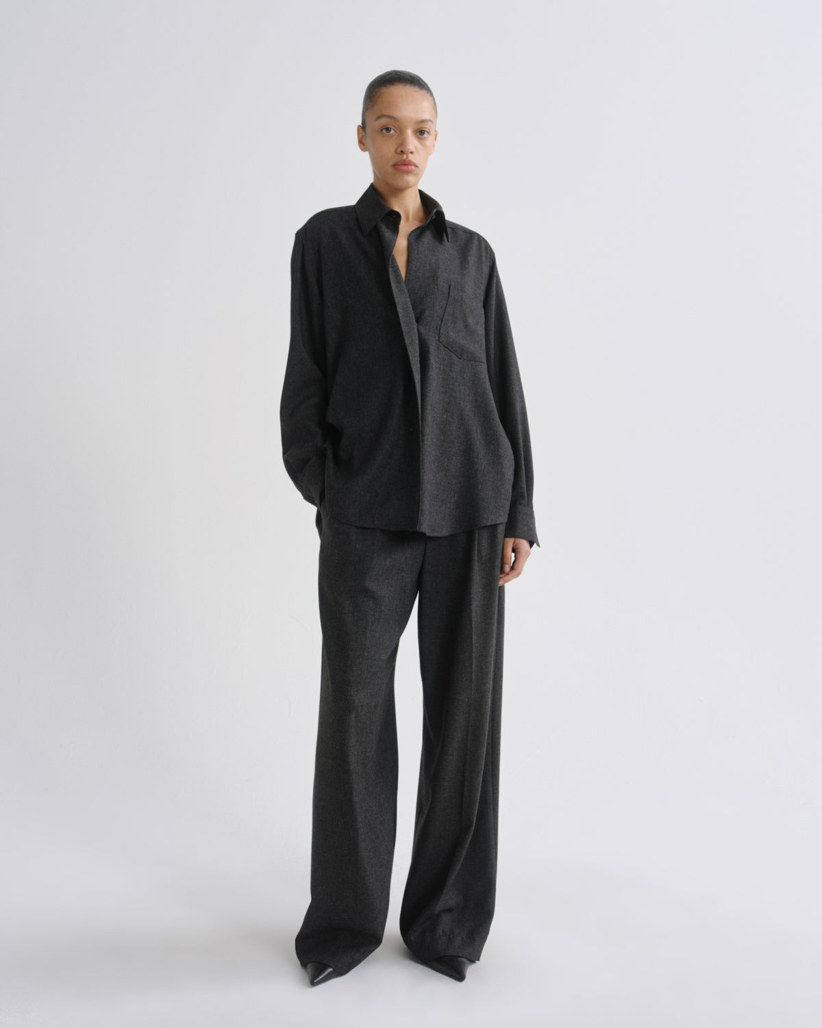 A model stands against a grey background wearing a dark charcoal grey two-piece linen set consisting of a long-sleeve button-down shirt and matching wide-leg trousers.