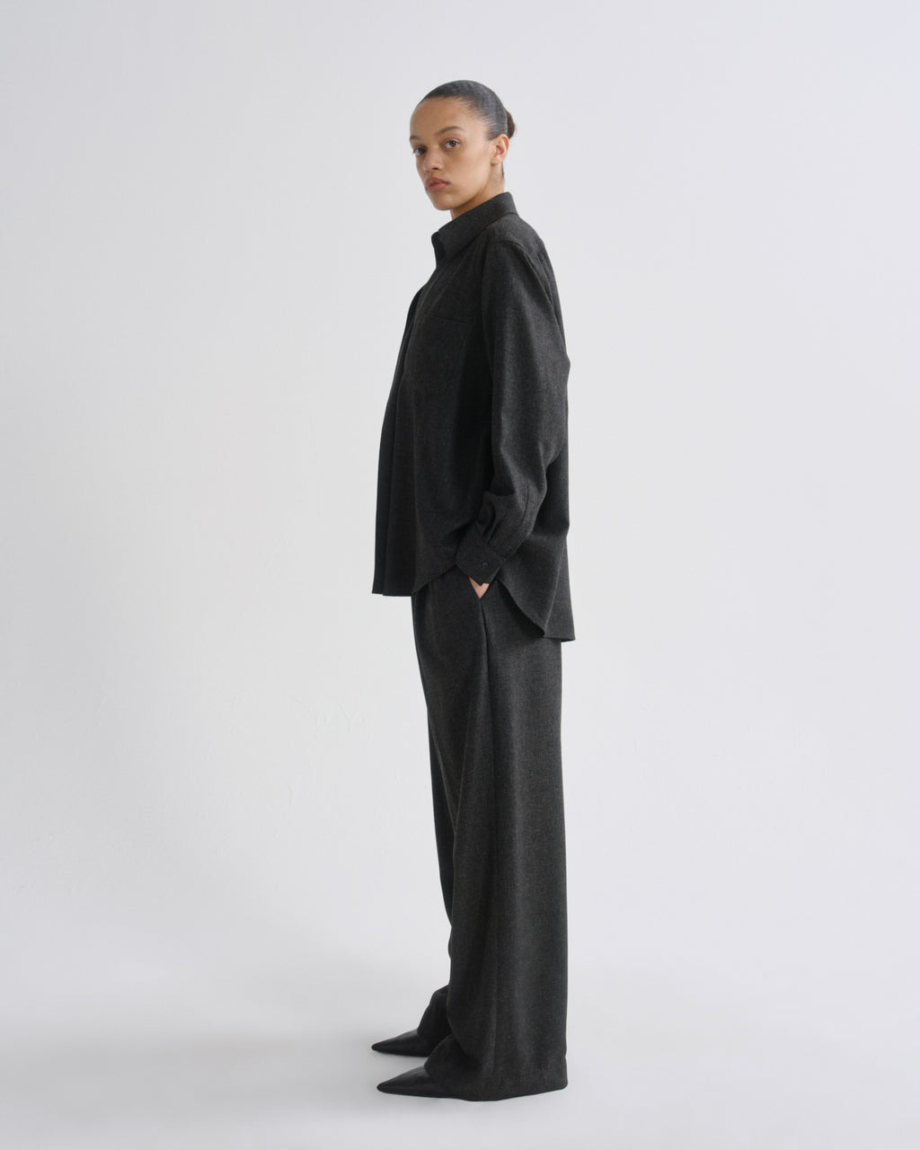 A model stands in profile wearing a dark charcoal grey two-piece outfit consisting of a high-neck, oversized long-sleeve top and matching wide-leg trousers,  against a grey background.