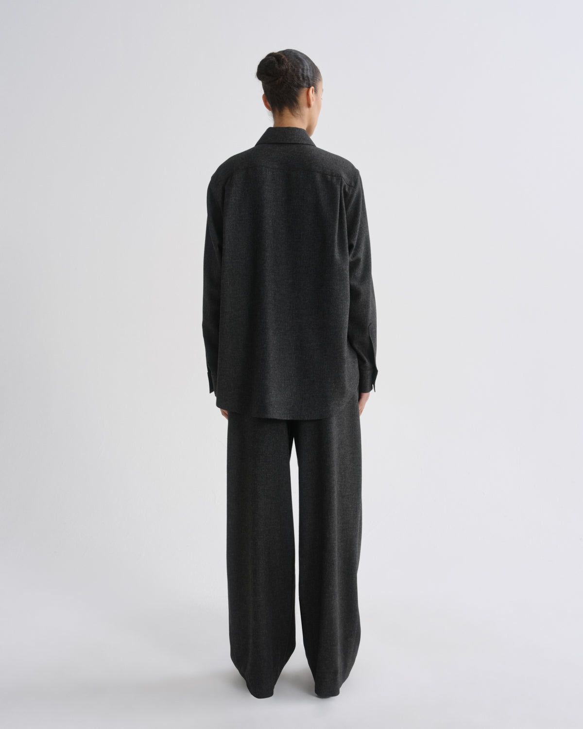 A model viewed from the back wearing a dark charcoal grey oversized long-sleeve shirt and matching wide-leg trousers against a grey background.