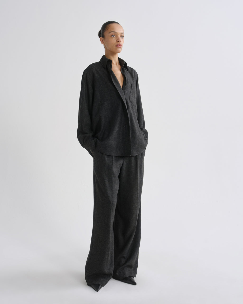 A model stands in a studio wearing a dark charcoal grey two-piece suit consisting of a long-sleeve button-up shirt and matching wide-leg trousers.