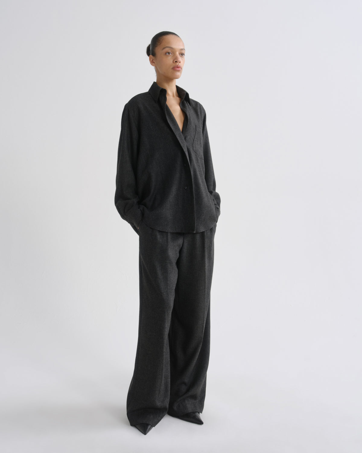 A model stands in a studio wearing a dark charcoal grey two-piece suit consisting of a long-sleeve button-up shirt and matching wide-leg trousers.
