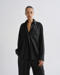 A model wearing a dark charcoal grey oversized button-down shirt and matching dark trousers, standing against a grey background.