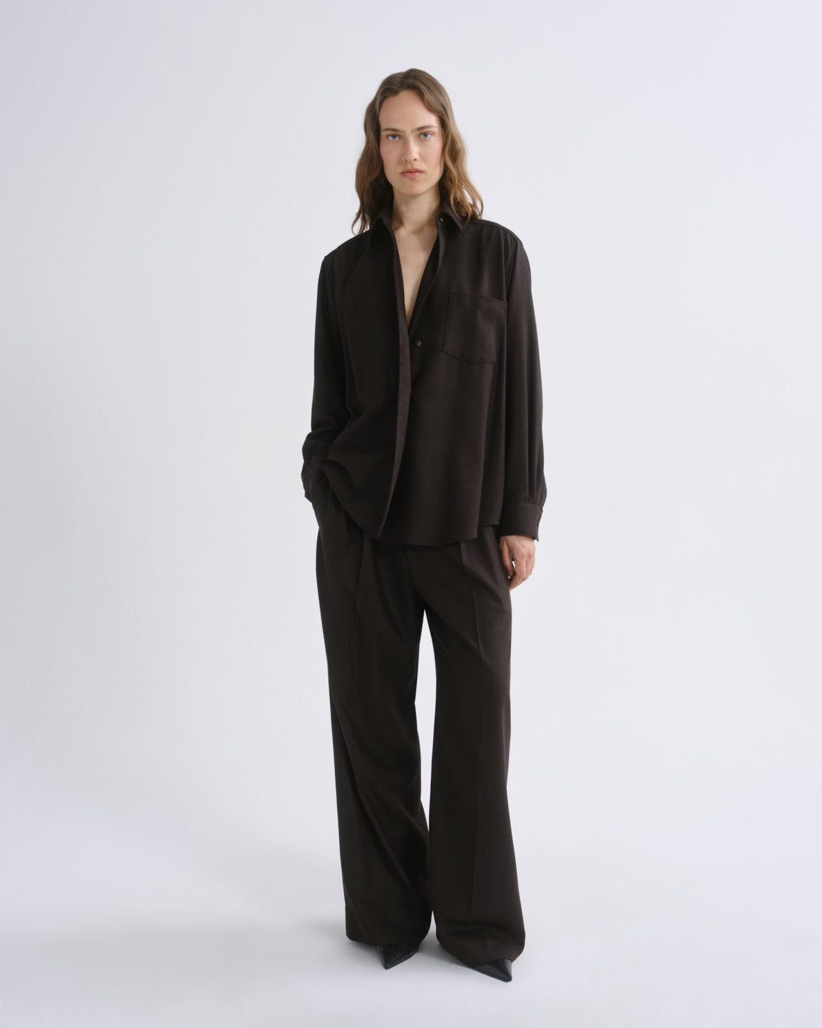 A model stands against a grey background wearing a dark brown long-sleeve button-front jacket with a relaxed, oversized fit and matching wide-leg trousers.