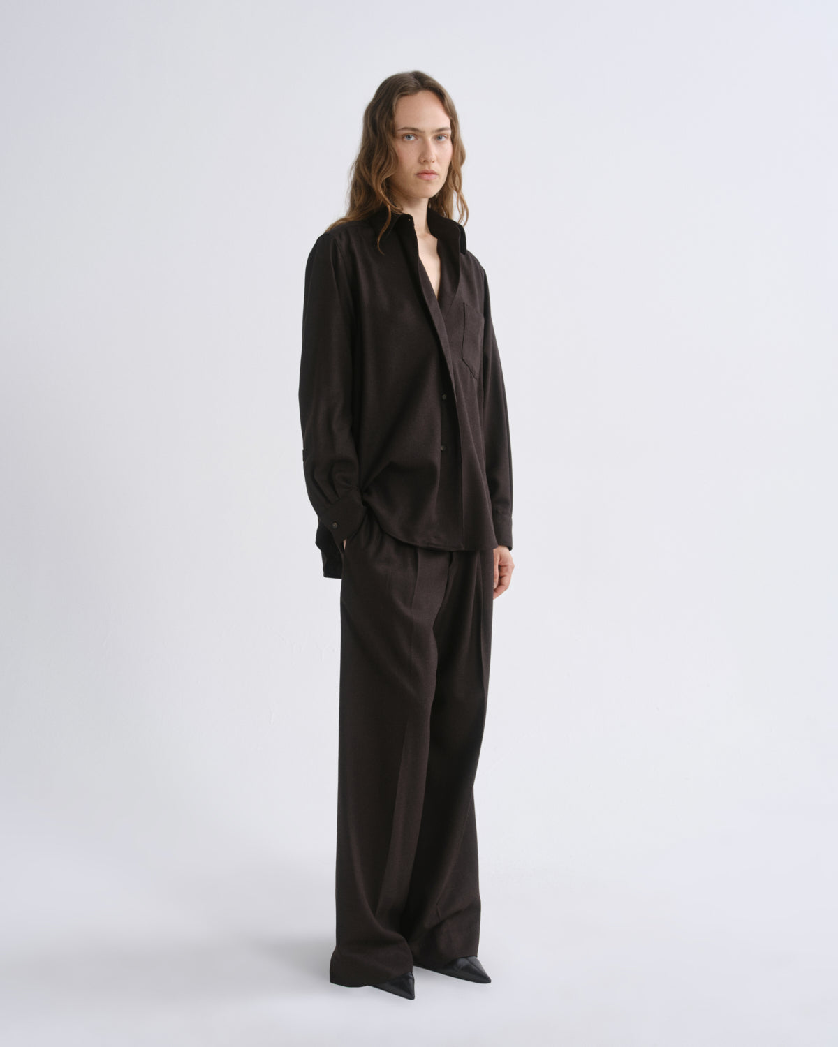 A model stands in a studio wearing a dark brown long-sleeve button-up shirt and matching wide-leg trousers against a grey background.
