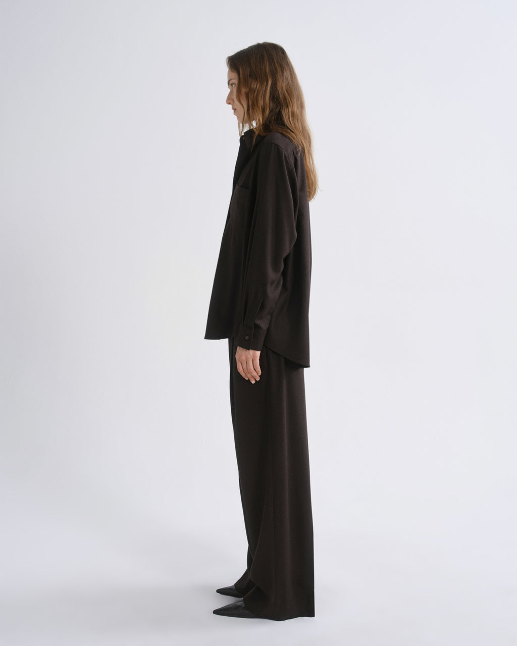 A model stands in profile wearing a black satin-like long-sleeve top and matching wide-leg trousers against a grey background.