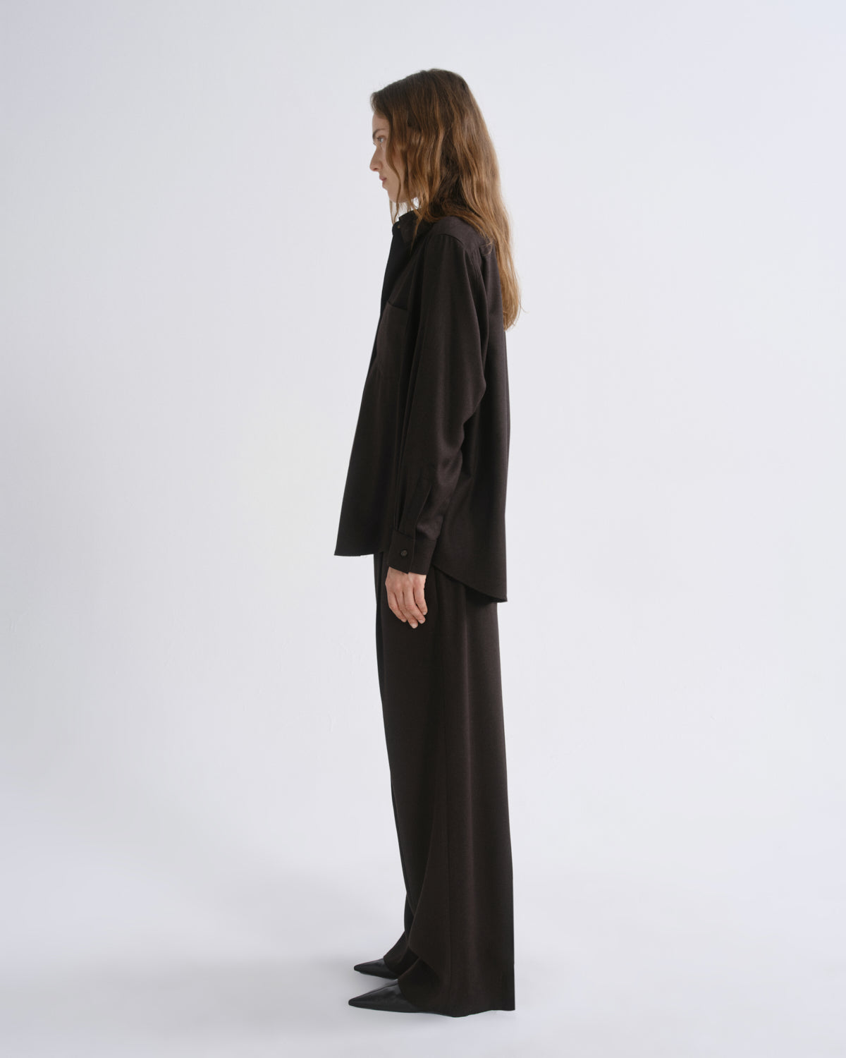 A model stands in profile wearing a black satin-like long-sleeve top and matching wide-leg trousers against a grey background.
