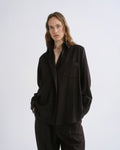 A woman modeling a loose-fitting black button-up shirt and matching black trousers against a grey background.