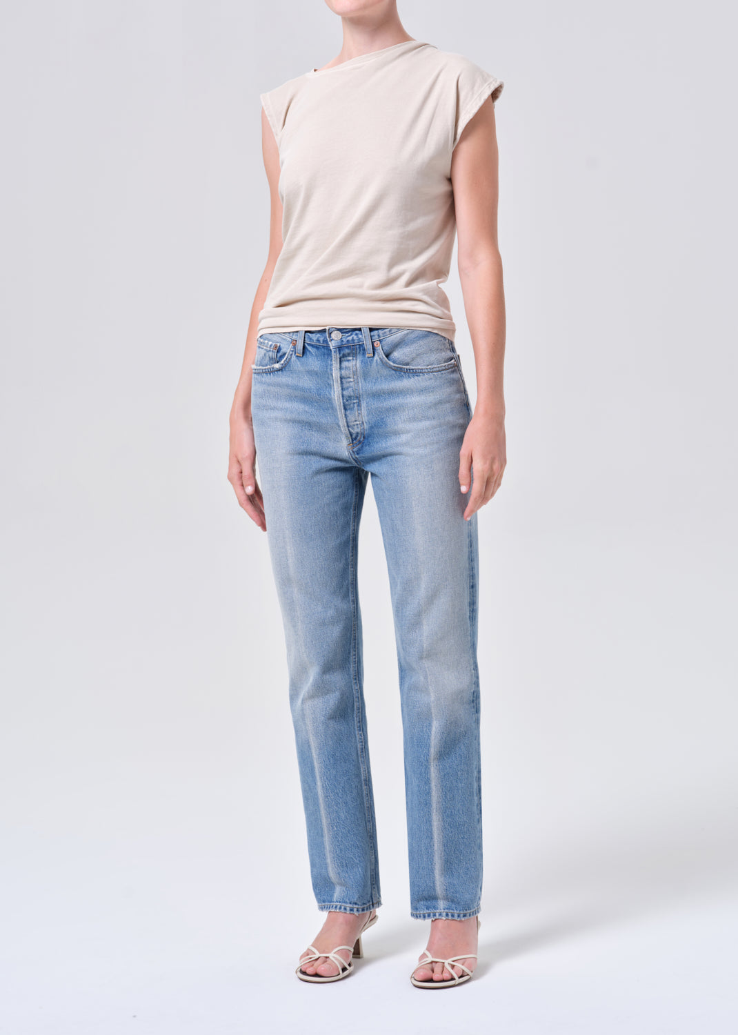 Person wearing a beige sleeveless top and light blue jeans on a white background