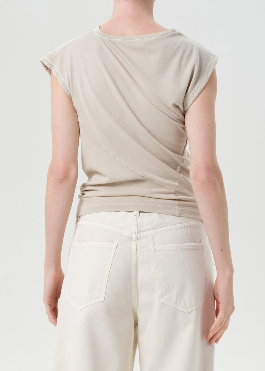 Person wearing a beige sleeveless top and white pants on a plain background