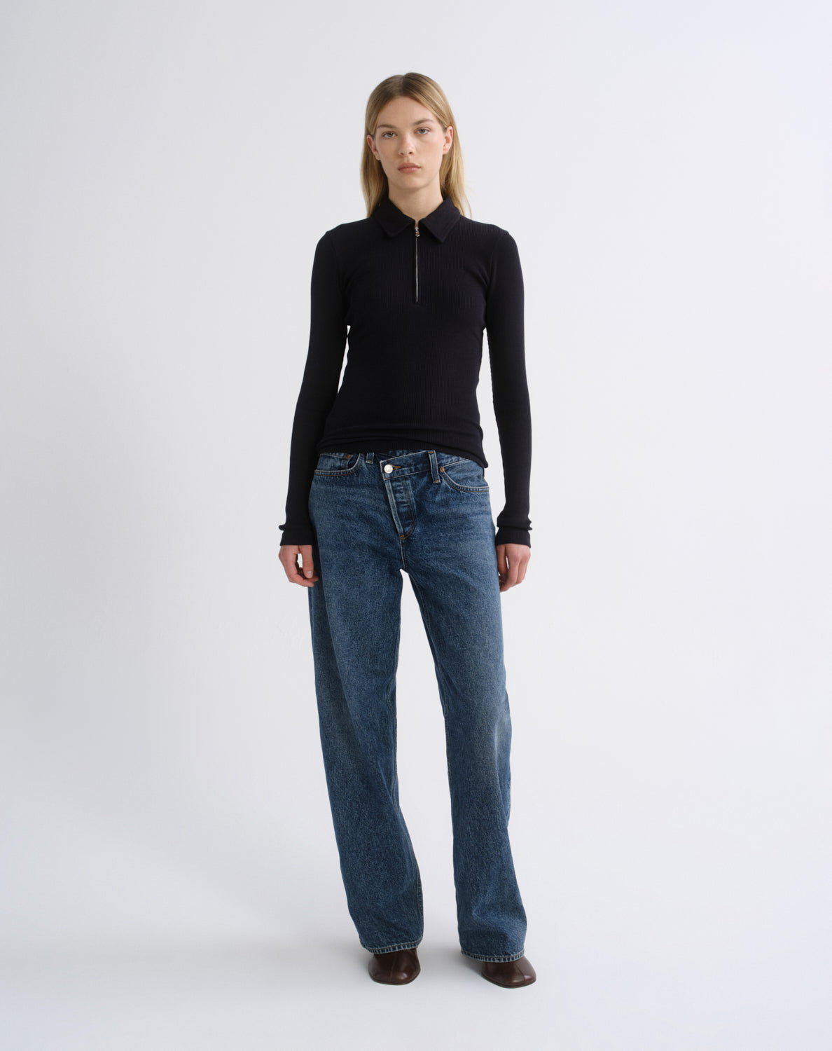 A woman stands against a light gray studio background wearing a fitted black long-sleeve zip-neck top, loose blue jeans, and dark brown shoes.