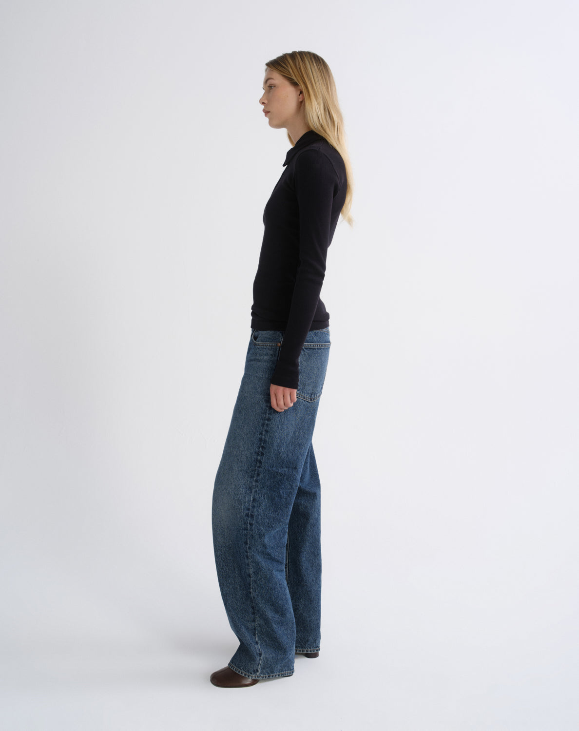 A woman standing in profile wearing a black long-sleeve top and wide-leg blue jeans against a plain light gray studio background.