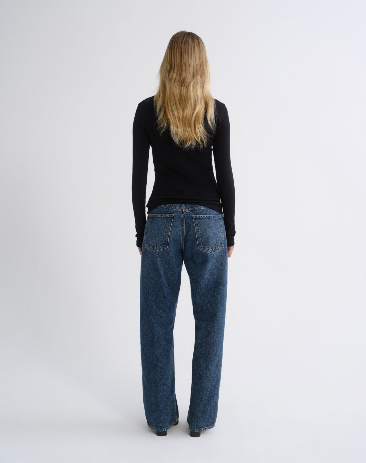 A woman viewed from behind wearing a black long-sleeve top and wide-leg blue jeans against a plain light gray studio background.