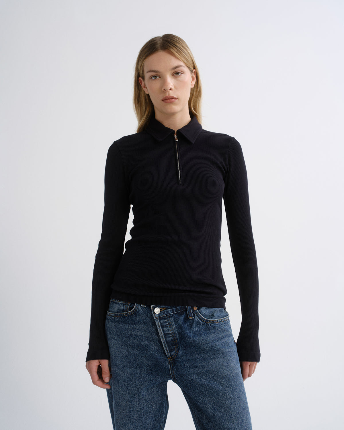 A woman modeling a black long-sleeve quarter-zip polo shirt with a collar, paired with blue jeans, against a plain light gray studio background.