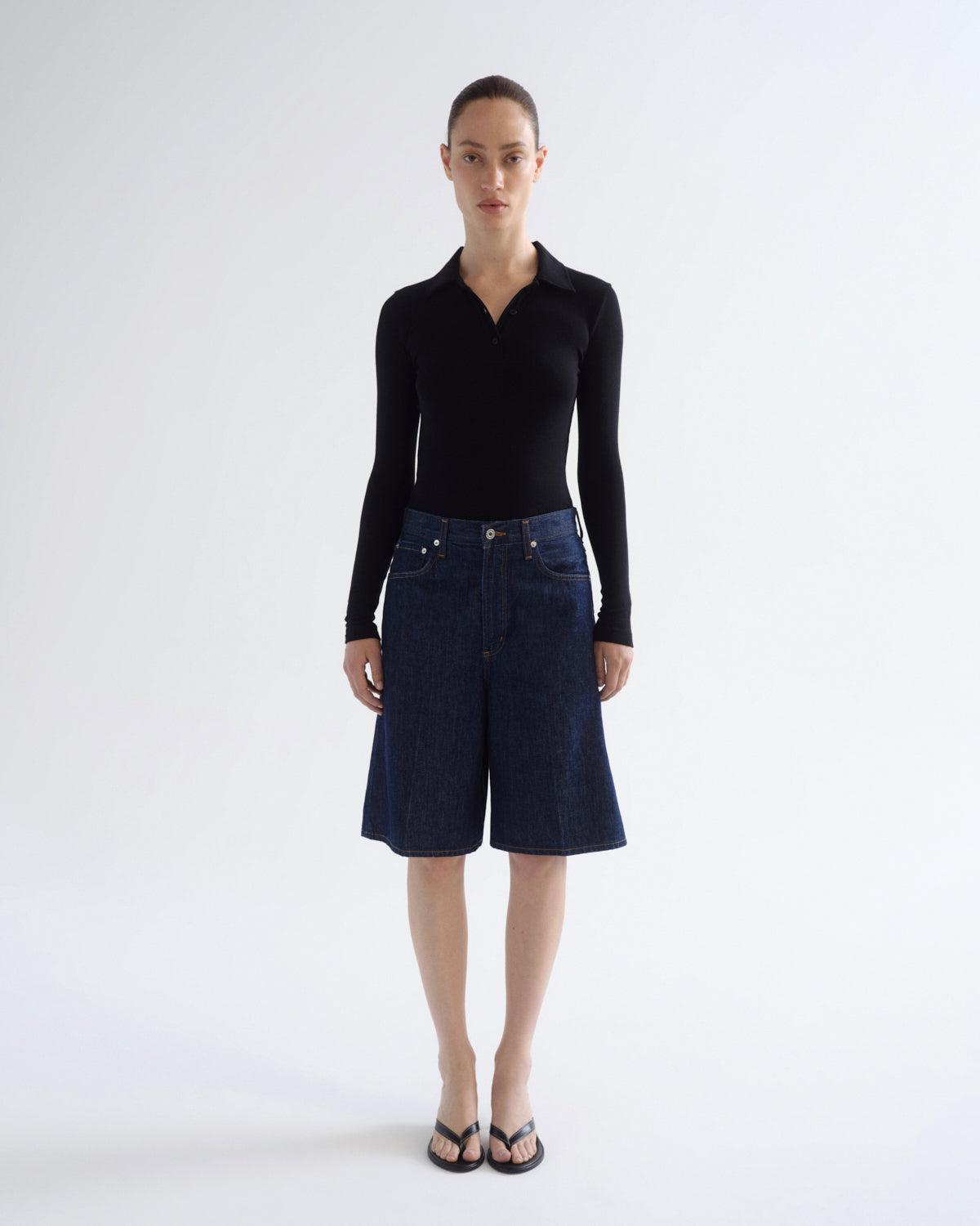 A model stands against a light gray studio background wearing a fitted black long-sleeve collared top, dark blue denim shorts, and black flip-flop sandals.