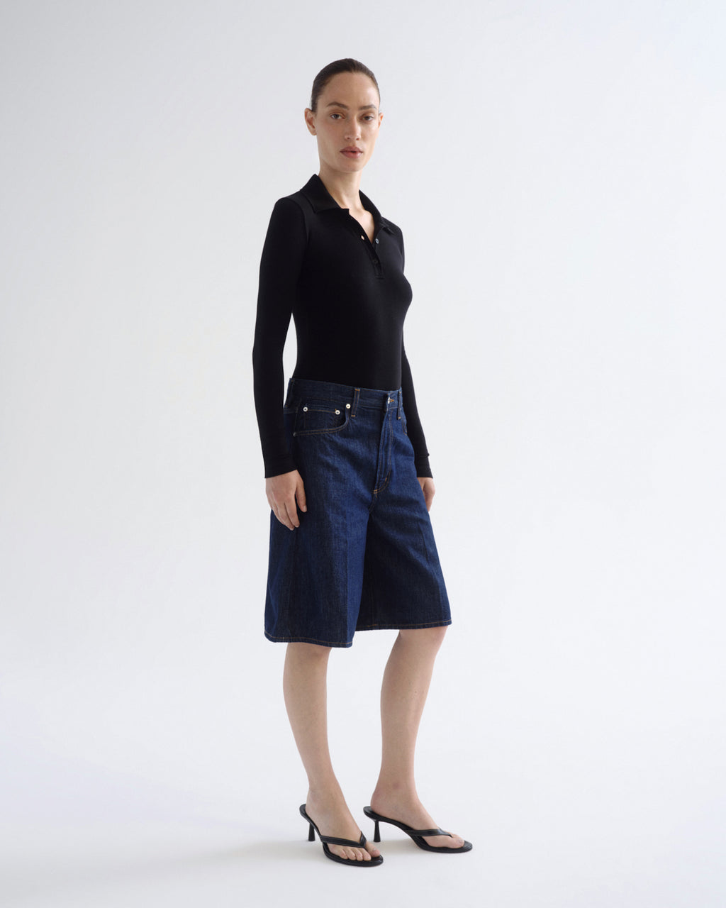 A model stands in a studio wearing a black long-sleeve polo shirt and dark blue denim shorts, paired with black strappy heels, against a plain light gray background.
