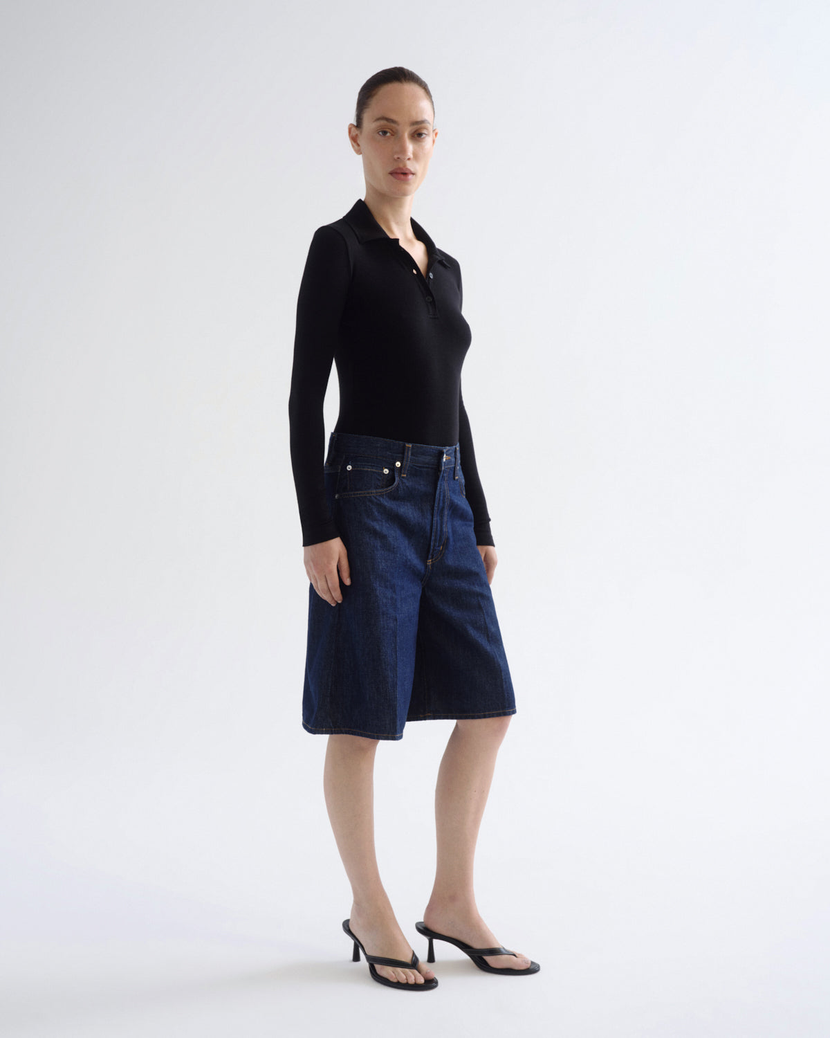 A model stands in a studio wearing a black long-sleeve polo shirt and dark blue denim shorts, paired with black strappy heels, against a plain light gray background.