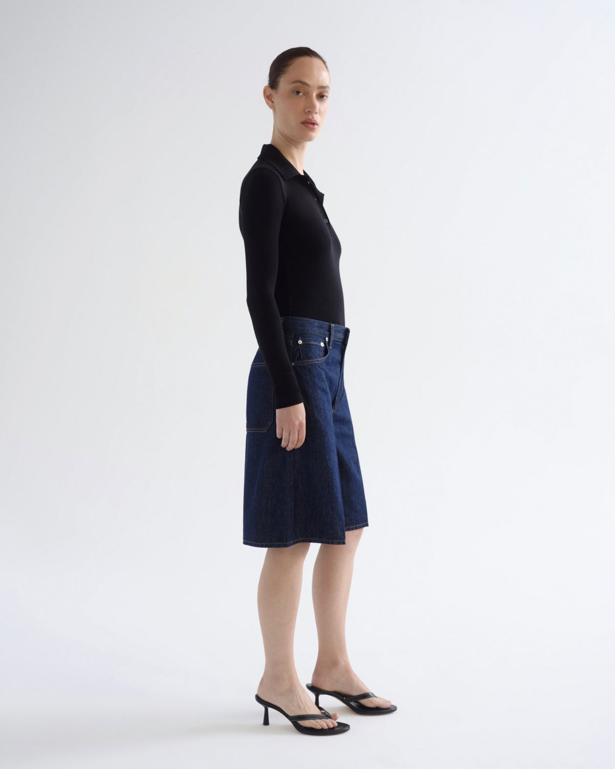 A model stands in profile wearing a black long-sleeve polo shirt and a dark blue denim midi skirt with front patch pockets, paired with black heeled sandals, against a plain white studio background.