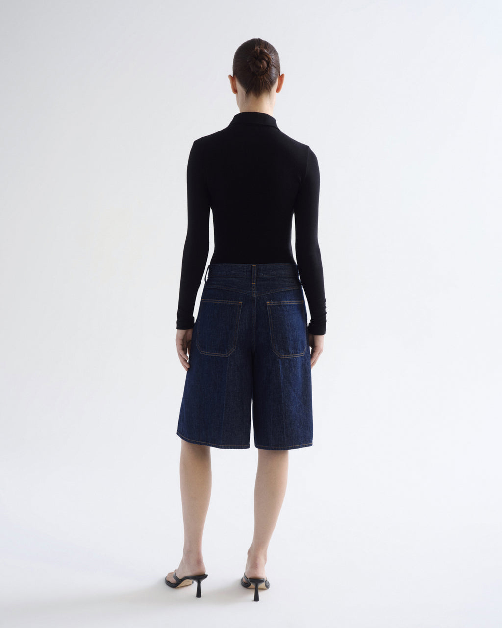 A model viewed from the back wearing a black long-sleeve mock neck top and dark blue denim shorts, paired with black high-heeled sandals, against a plain white studio background.