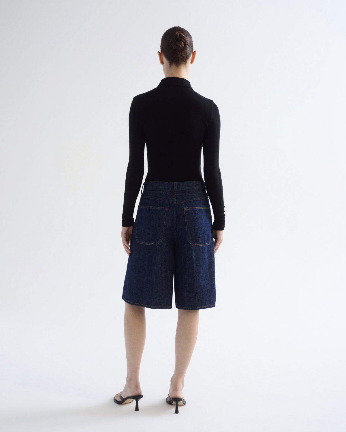 A model viewed from the back wearing a black long-sleeve mock neck top and dark blue denim shorts, paired with black high-heeled sandals, against a plain white studio background.