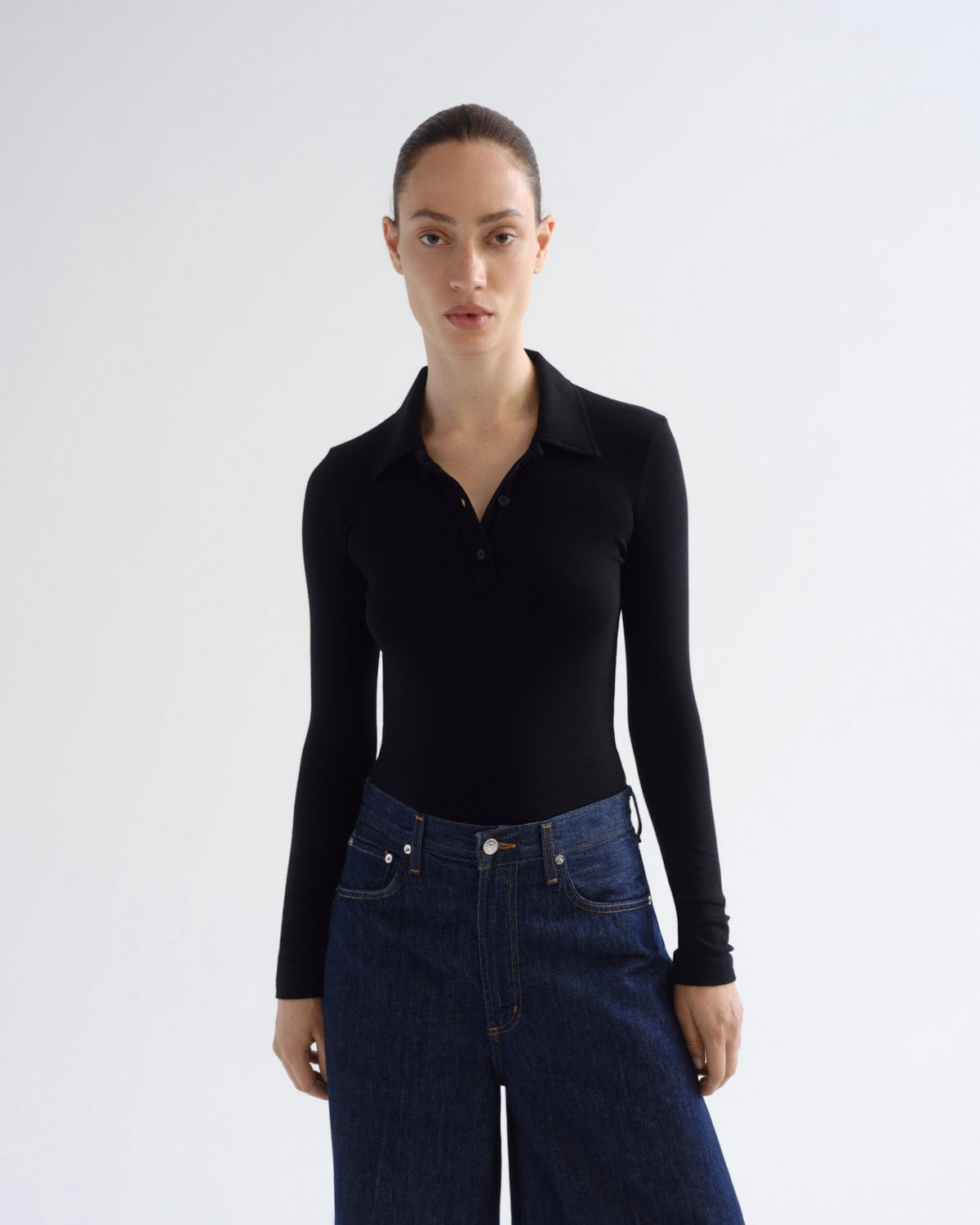 A woman modeling a black long-sleeve polo shirt tucked into dark blue jeans, standing against a plain light gray studio background.