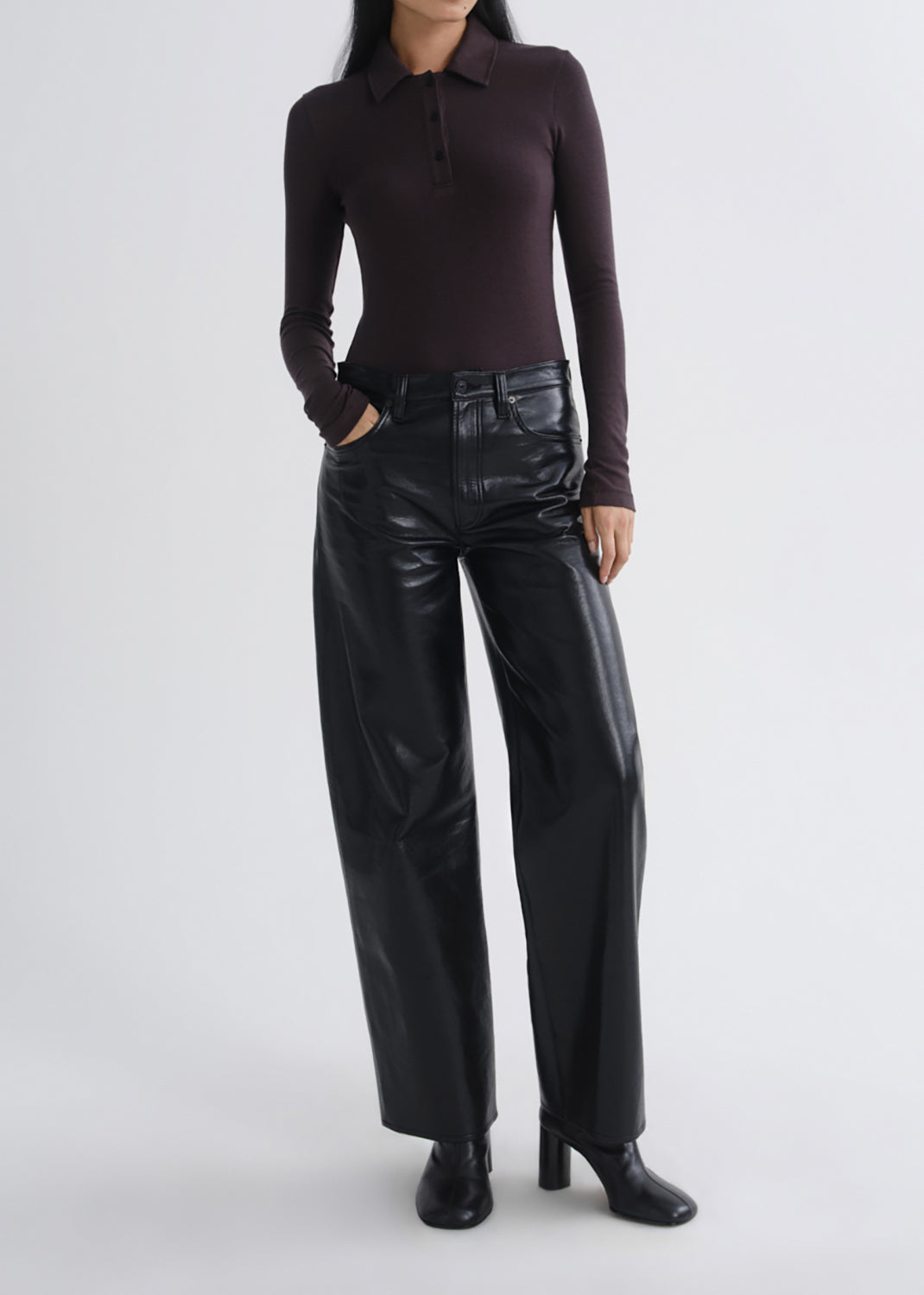 A model wearing a dark brown long-sleeve polo shirt and shiny black wide-leg trousers, standing against a grey background.