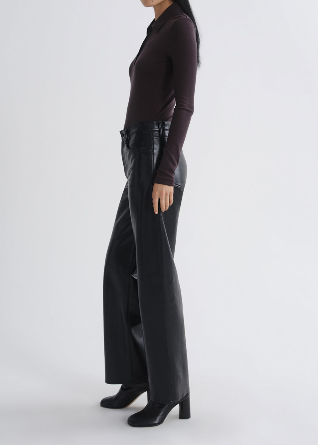 A model shown from the side wearing a dark brown long-sleeve collared top, black wide-leg leather pants, and black heeled ankle boots against a grey background.