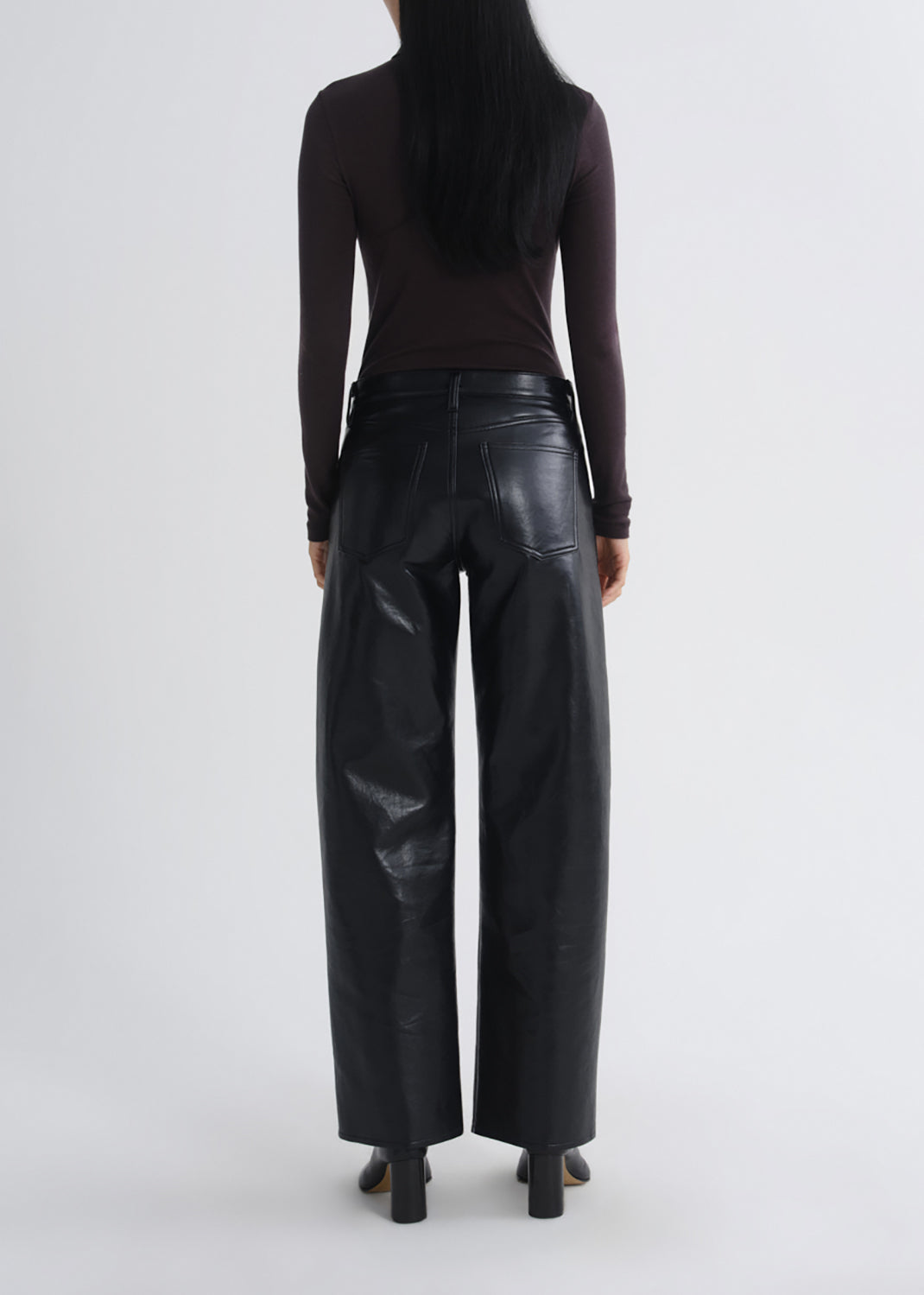 A model shown from the back wearing black wide-leg faux leather pants, a dark brown long-sleeve top, and black heels against a grey background.